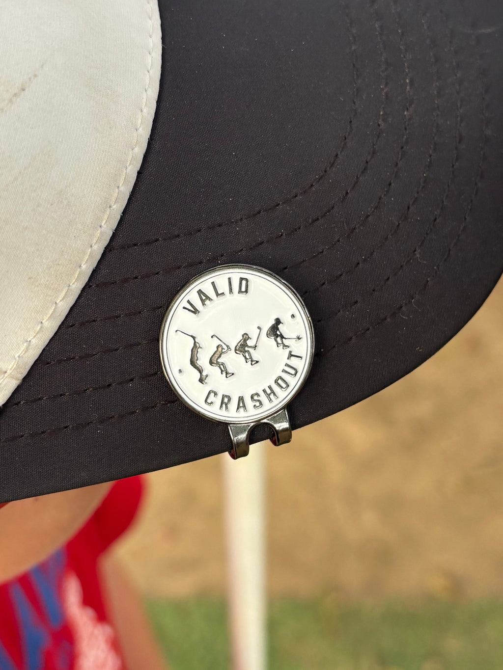 A close-up of a golf hat featuring a metal golf ball marker that reads 'VALID CRASHOUT' 